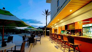 An outdoor bar area with seating and umbrellas, a pool in the background, and a building with a red facade. - The Lucie (Jensen Beach)