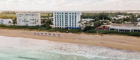 Un hotel frente a la playa con varios pisos, una playa arenosa con sombrillas y vistas al océano.