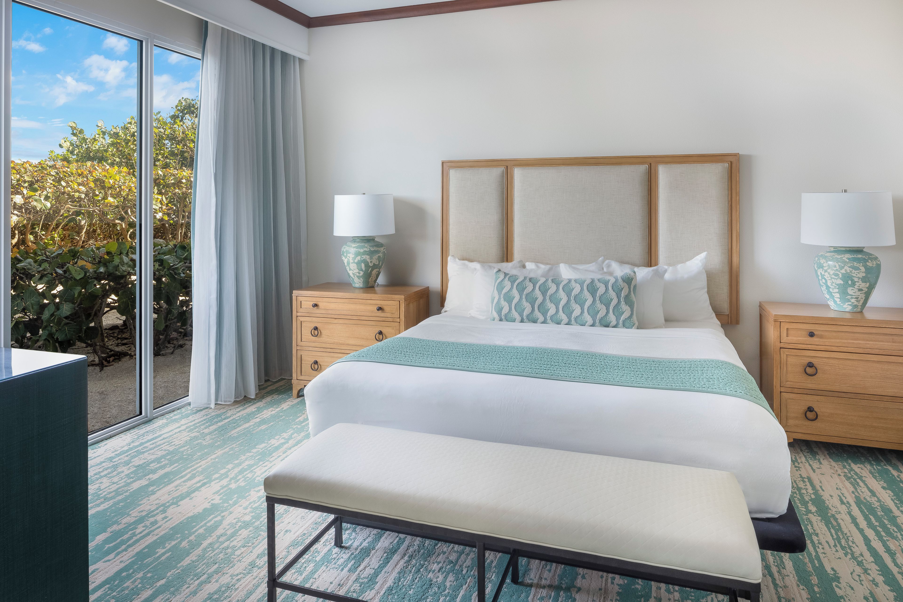 Une chambre d’hôtel avec un grand lit, deux tables de chevet avec des lampes, un banc, une vue sur la verdure par la fenêtre et une tête de lit en bois.