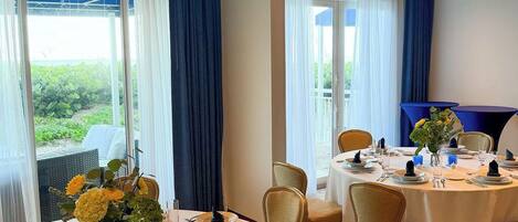 Uma sala de jantar de hotel com mesas redondas, cadeiras e vista para o verde lá fora.