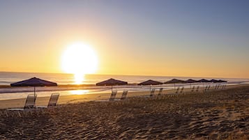 A beach with rows of sun loungers and umbrellas, a clear sky, and the sun setting over the ocean.