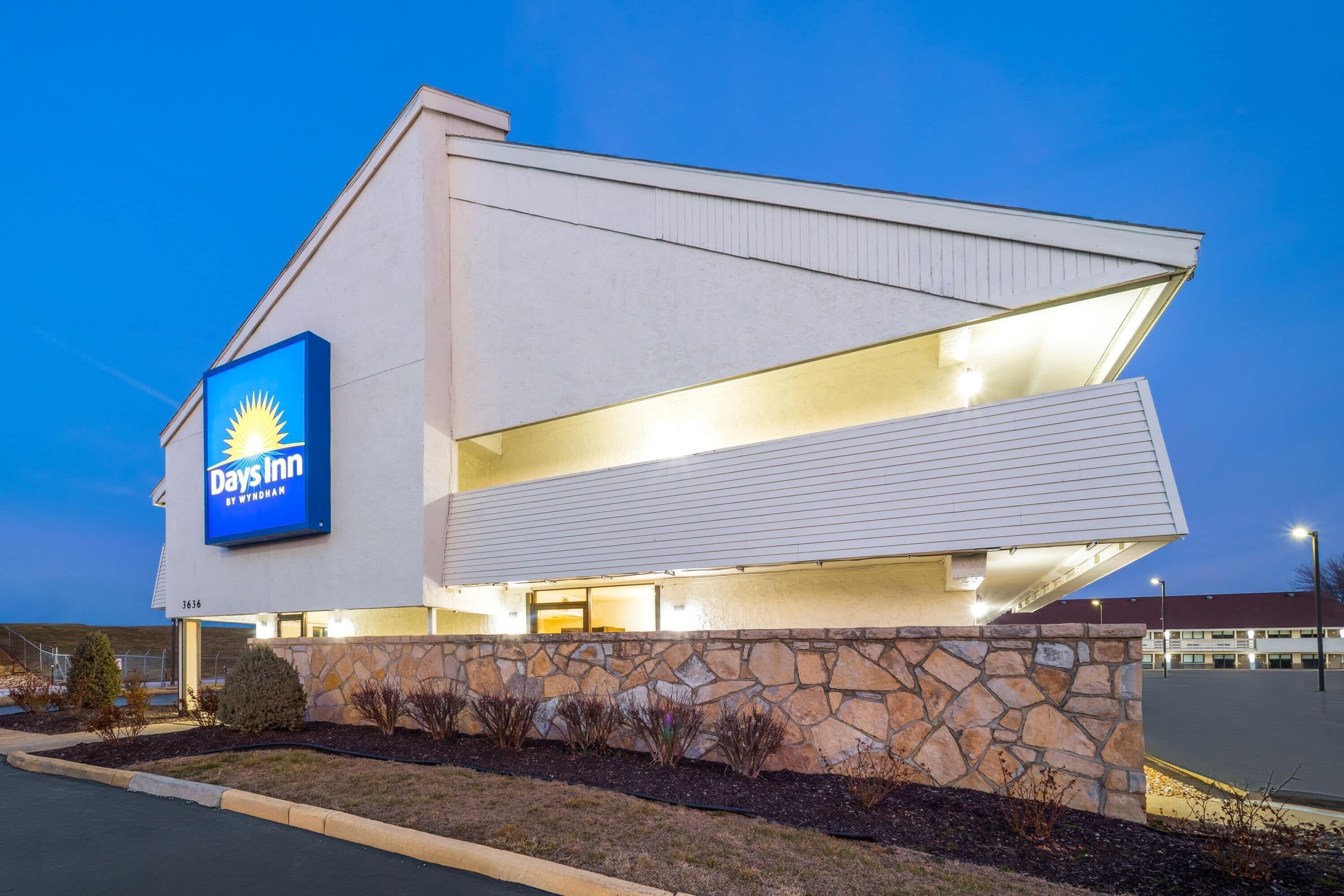 Un edificio Days Inn con fachada de piedra, un letrero iluminado grande y un estacionamiento al fondo.