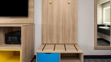Una habitación con un televisor montado sobre un mueble de madera, un armario de madera con dos ganchos, un pequeño archivador azul y un espejo que refleja un baño con lavabo y espejo.
