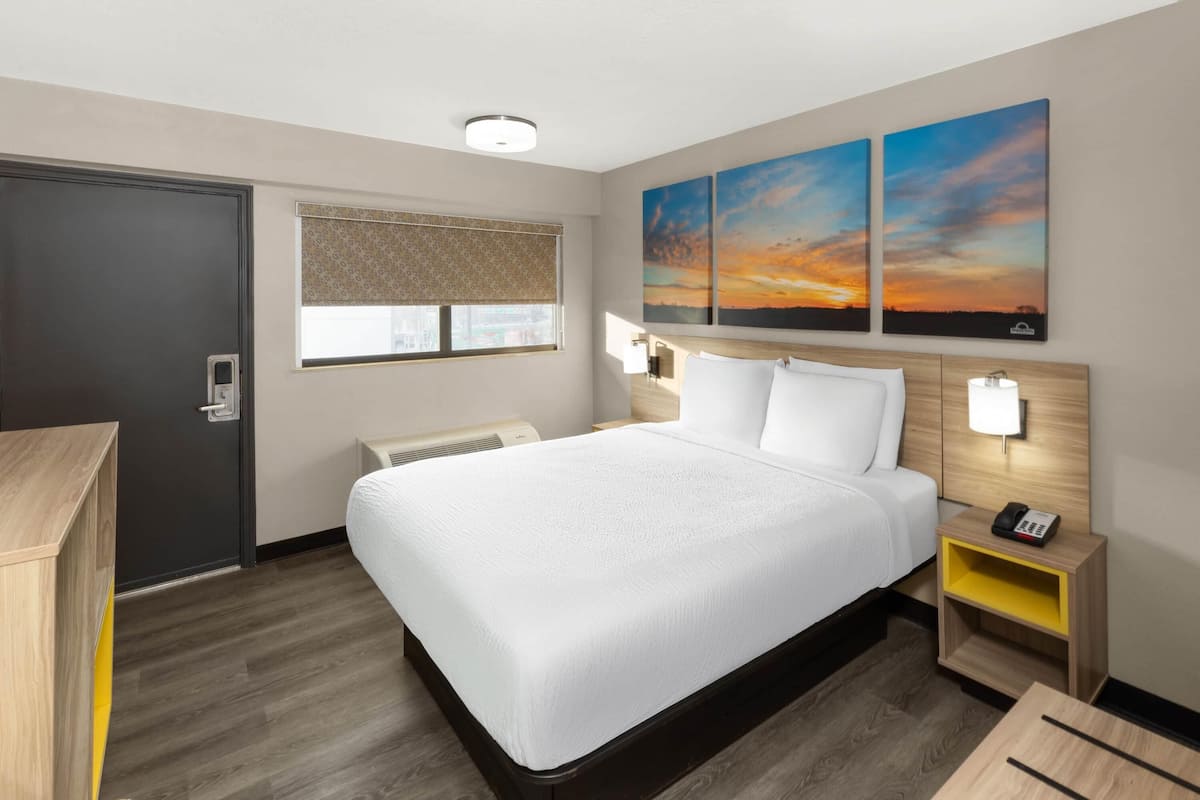 A hotel room with a large bed, a bedside table with a lamp, a wall-mounted phone, and a window with blinds.