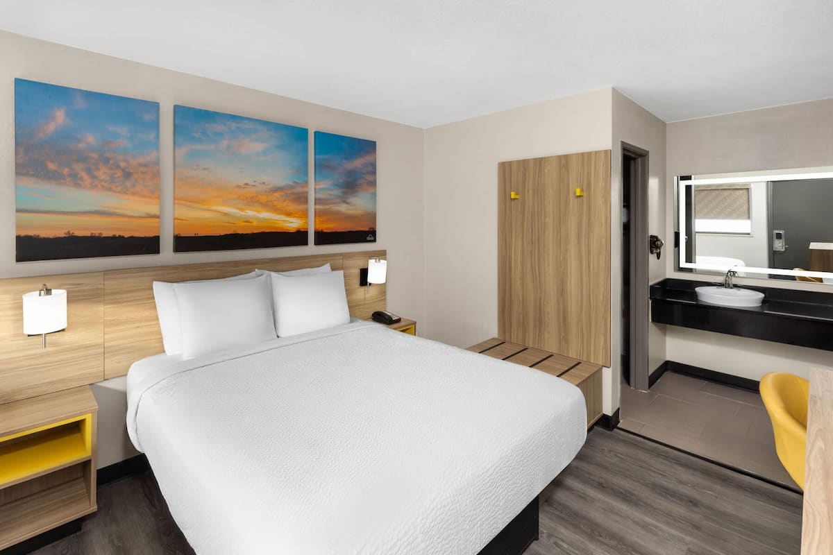 A hotel room with a large bed, a wooden wardrobe, a bathroom with a mirror, and two sunset paintings on the wall.