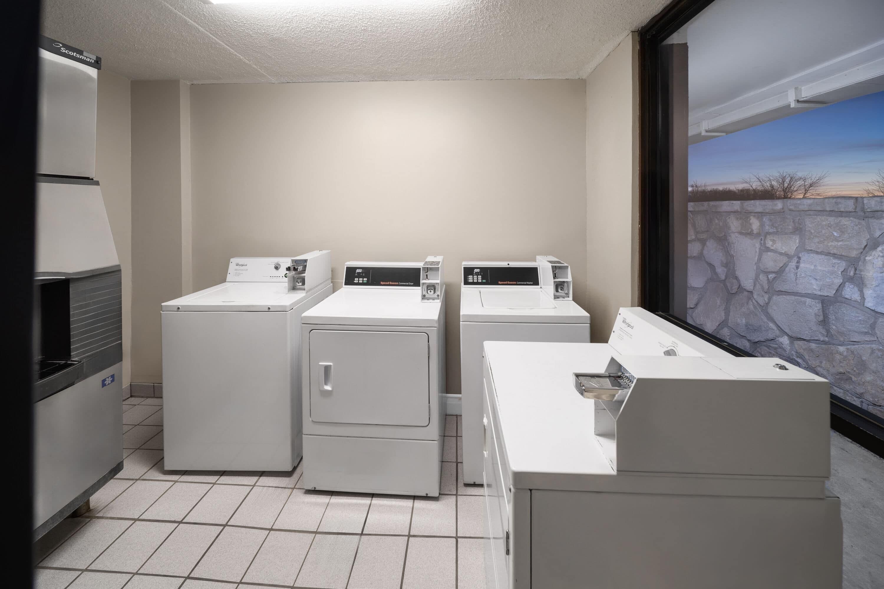 A laundry room with four white washers and dryers, a tiled floor, and a stone wall with a window.