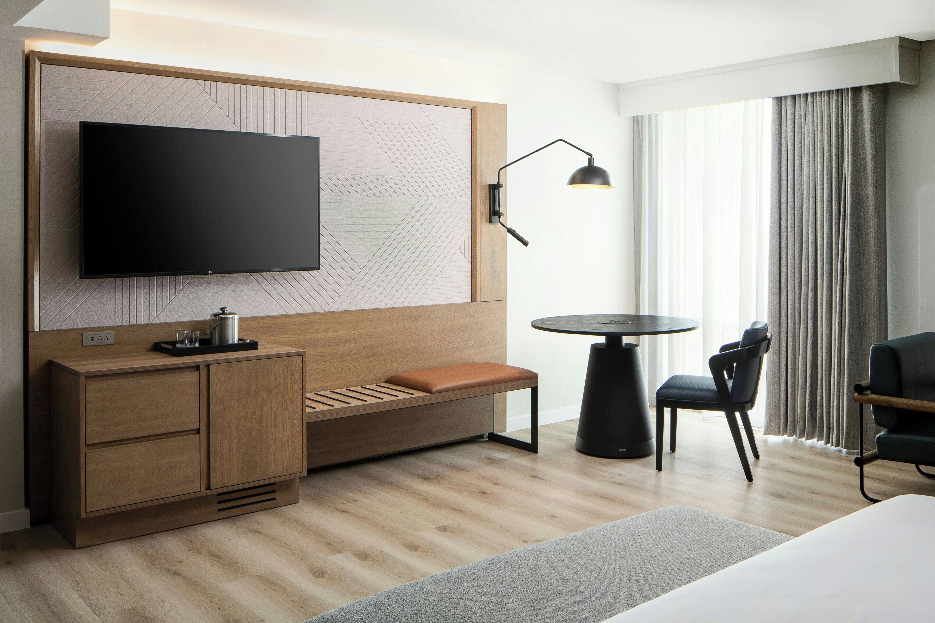 Habitación de hotel moderna con un televisor de pantalla plana grande montado en una pared de paneles de madera, un pequeño banco de madera con cojín, una mesa redonda negra con dos sillas y un amplio ventanal con cortinas.