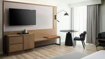 Habitación de hotel moderna con un televisor de pantalla plana grande montado en una pared de paneles de madera, un pequeño banco de madera con cojín, una mesa redonda negra con dos sillas y un amplio ventanal con cortinas.