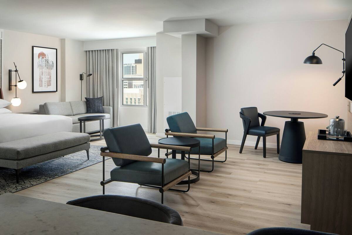Una habitación de hotel moderna con una cama, dos sillones, una mesita y un escritorio. Hay una ventana con cortinas, un cuadro en la pared y una lámpara sobre el escritorio.