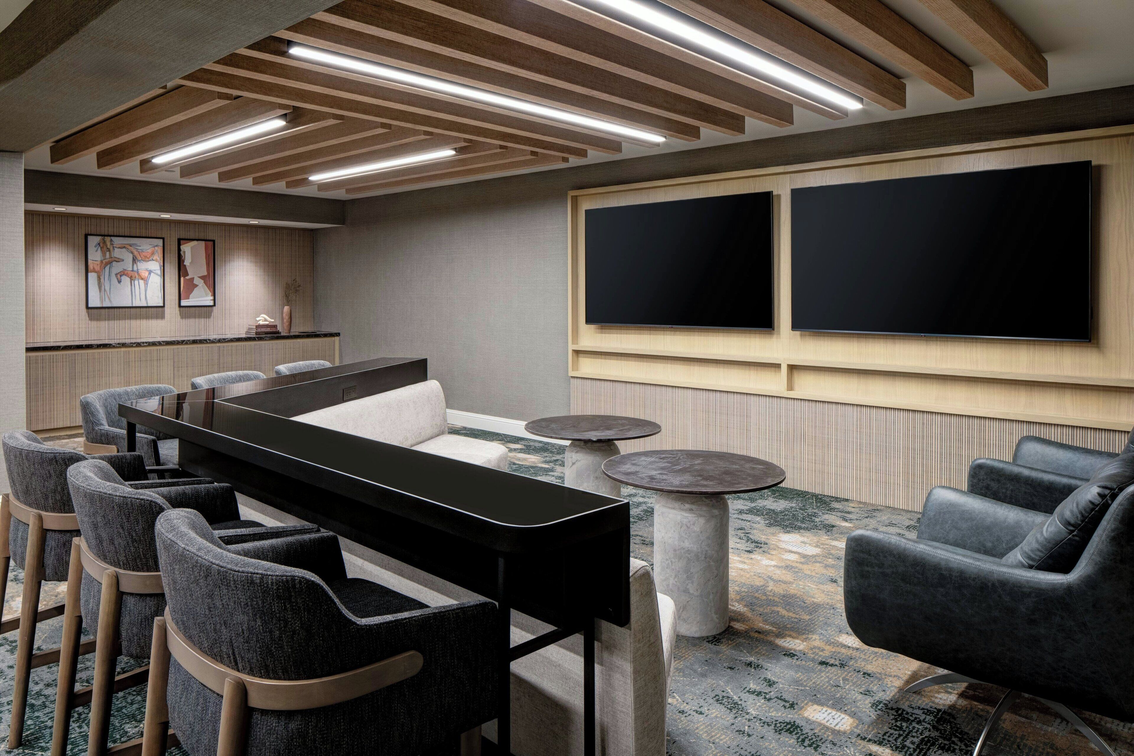A modern lounge area with a long table, multiple chairs, and two large screens on the wall.