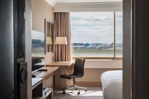 A hotel room with a desk, chair, and bed, a window with a view of an airport and planes.
