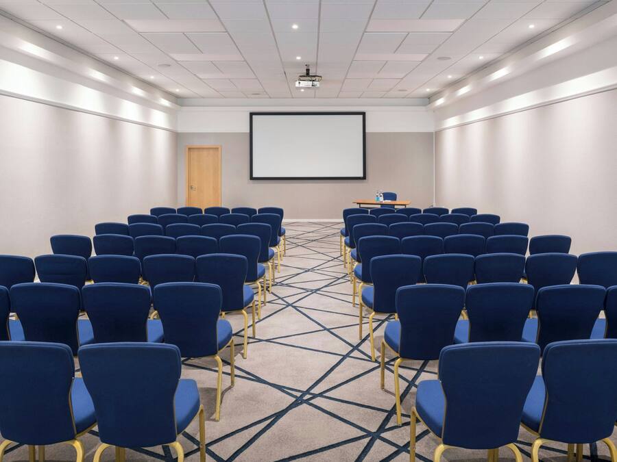Un salón de conferencias con filas de sillas azules, una pantalla de proyector y una alfombra con un diseño geométrico.