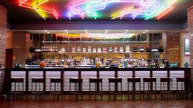 Un bar dal design moderno, con un soffitto illuminato da luci al neon e una vasta selezione di bottiglie e bicchieri sugli scaffali.
