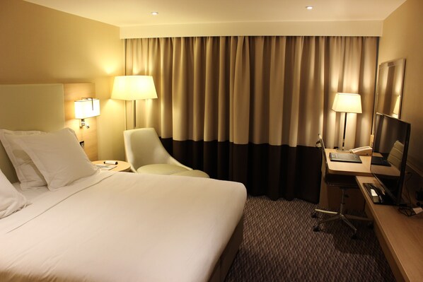 A hotel room with a large bed, two bedside tables with lamps, a desk with a chair, a flat-screen TV, and a large window with curtains. - Renaissance London Heathrow Hotel (Hounslow)