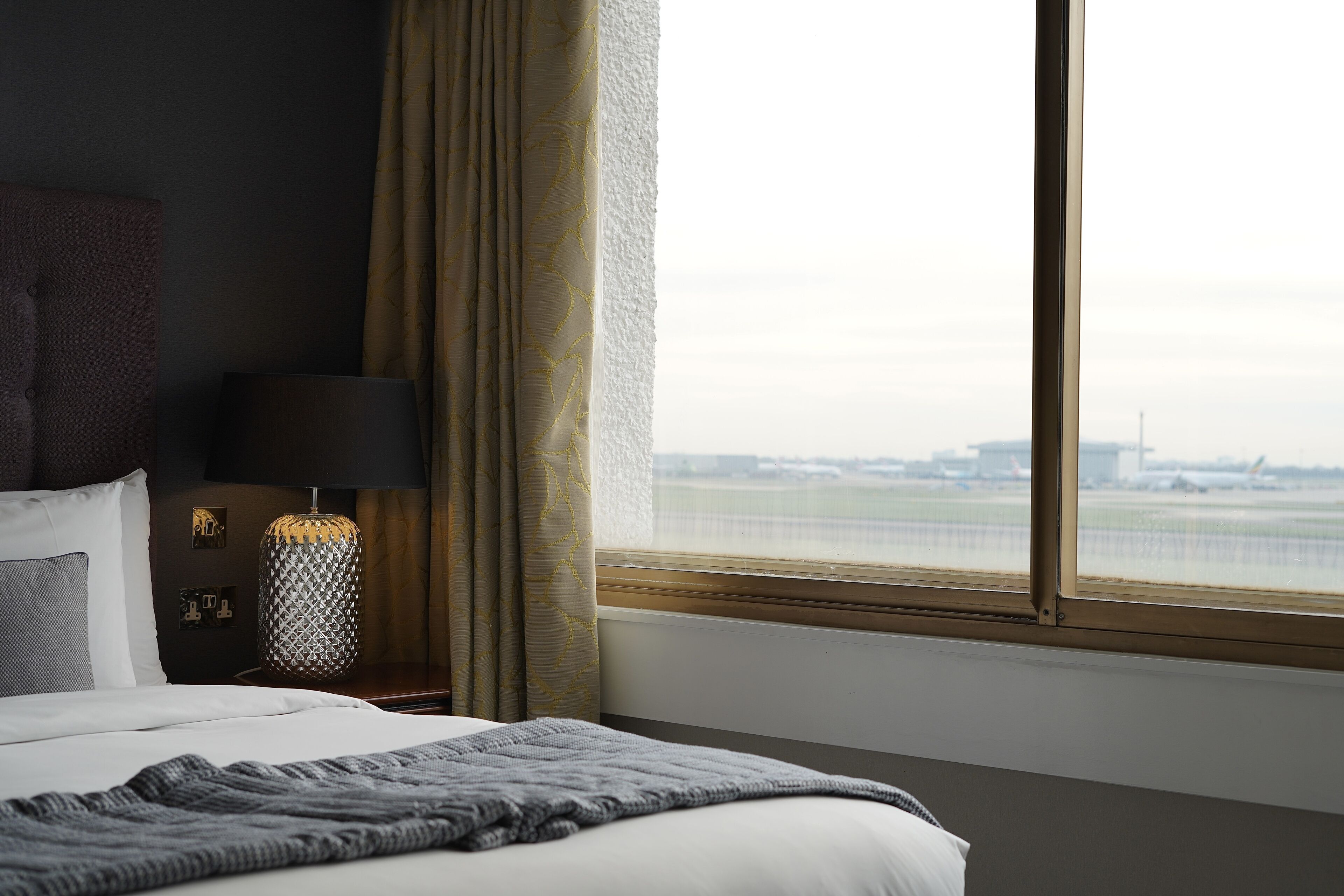 A hotel room with a bed, a nightstand with a lamp, and a view of an airport.