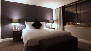 Una camera d'albergo moderna con un letto grande, due comodini con lampade, un poggiapiedi scuro e un'ampia finestra che si affaccia sulla città.