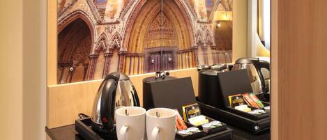 A hotel room with a large mirror reflecting a detailed mural of a cathedral. The room includes a black countertop with a kettle, two mugs, and a tray with breakfast items.