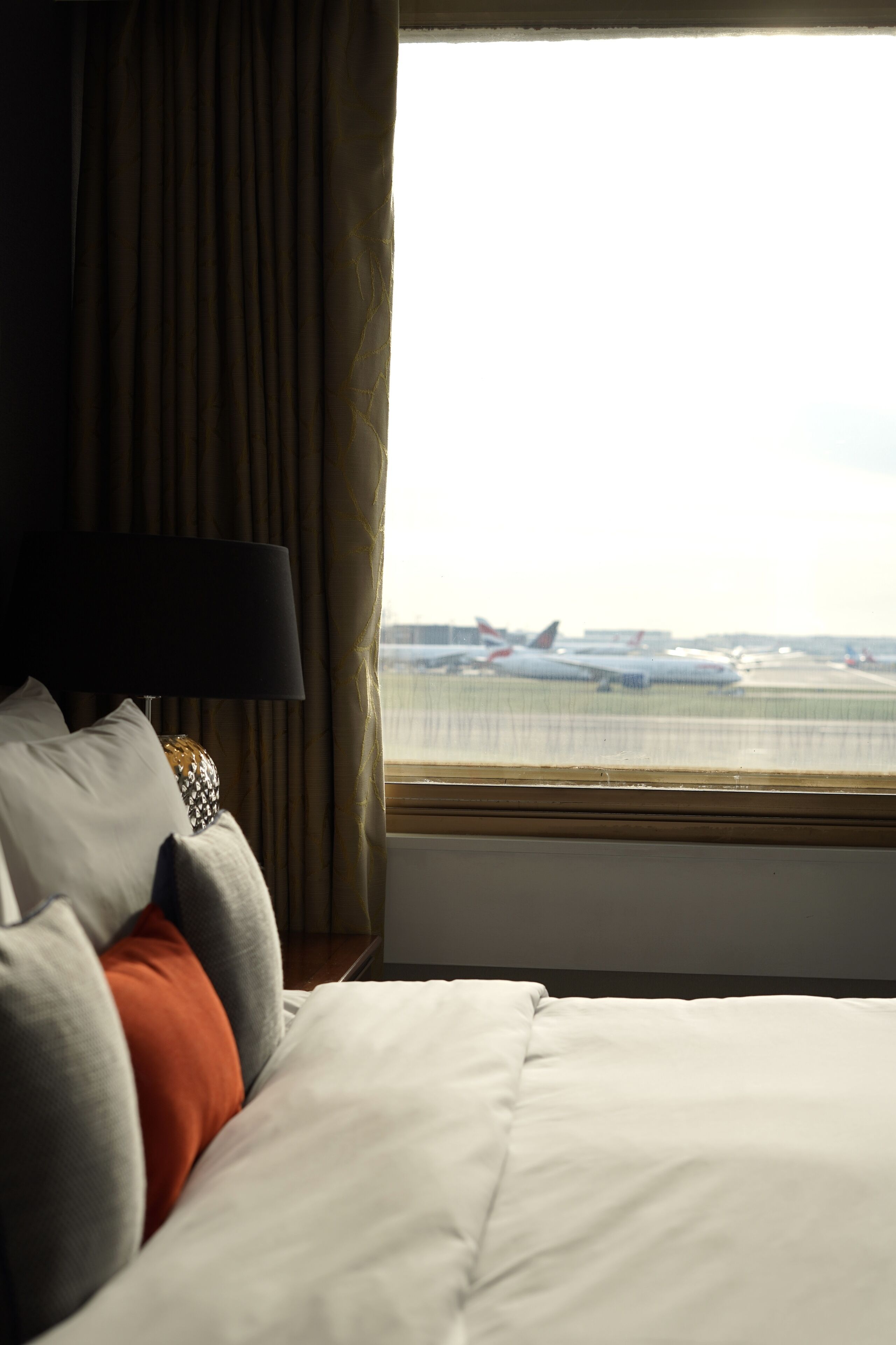 A hotel room with a bed, a lamp, and a view of airplanes on the tarmac through a window.