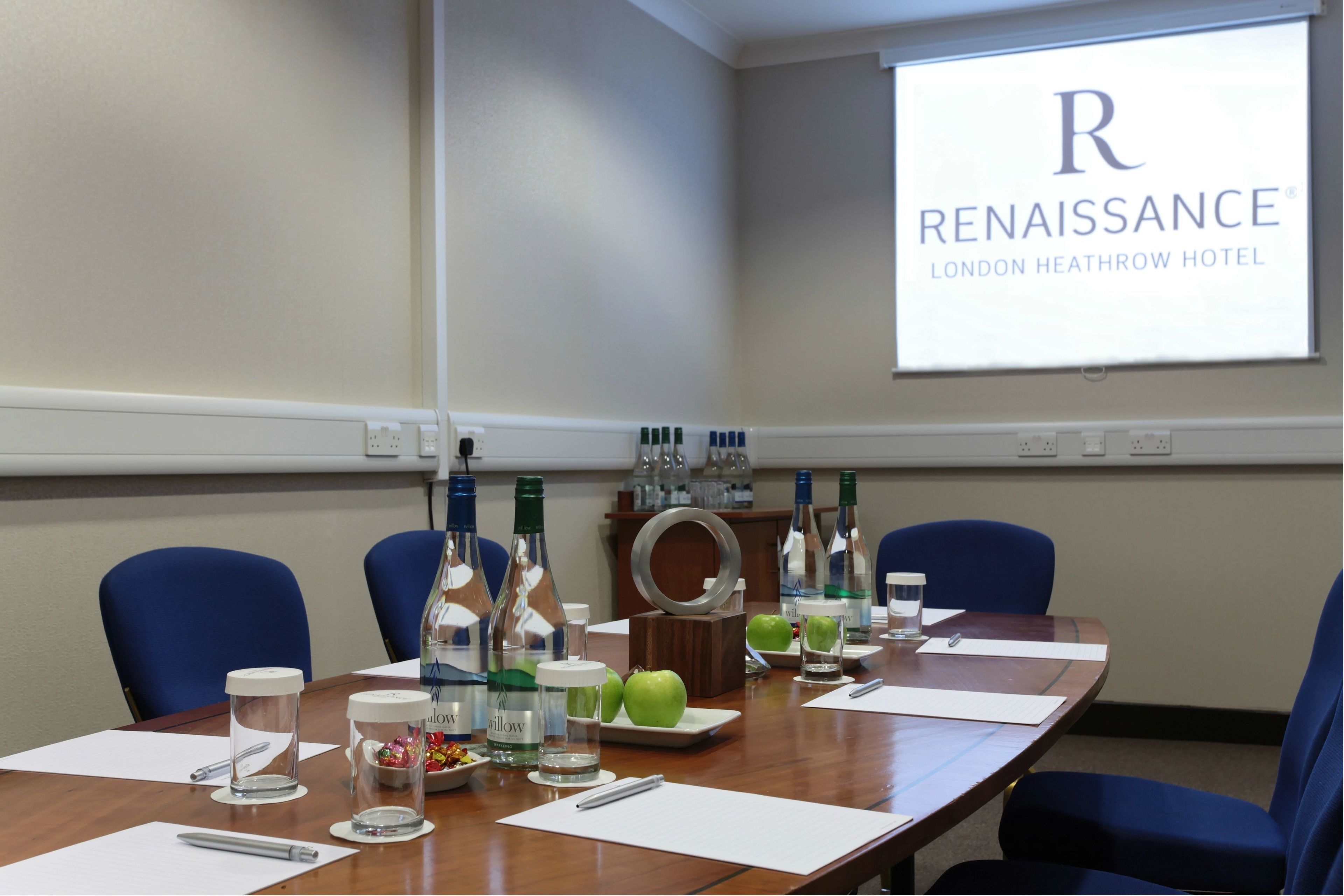 A conference room at the Renaissance London Heathrow Hotel with a long table, blue chairs, and various amenities like bottles, apples, and glasses.