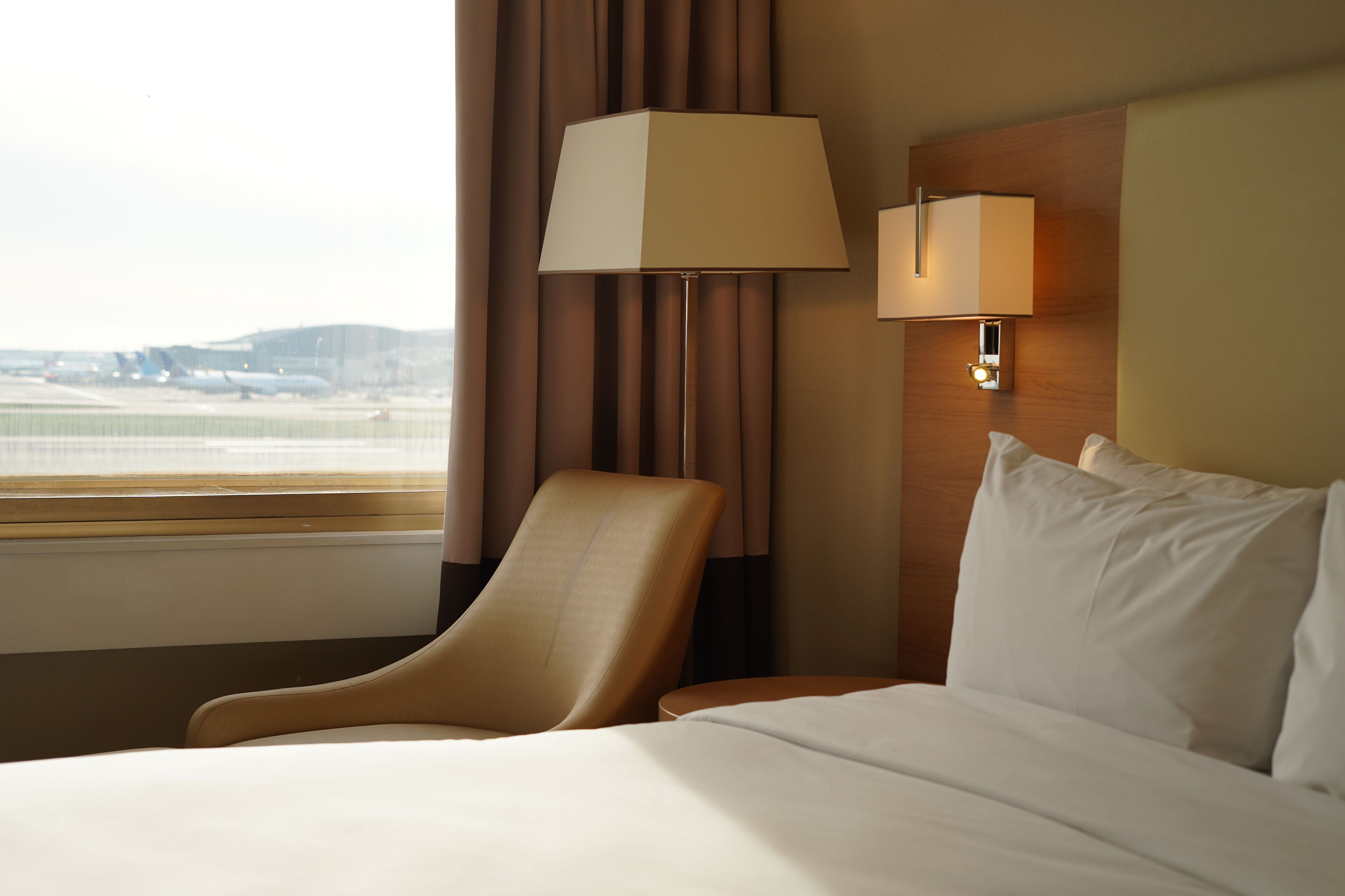A hotel room with a bed, a chair, a lamp, and a window with a view of the airport.