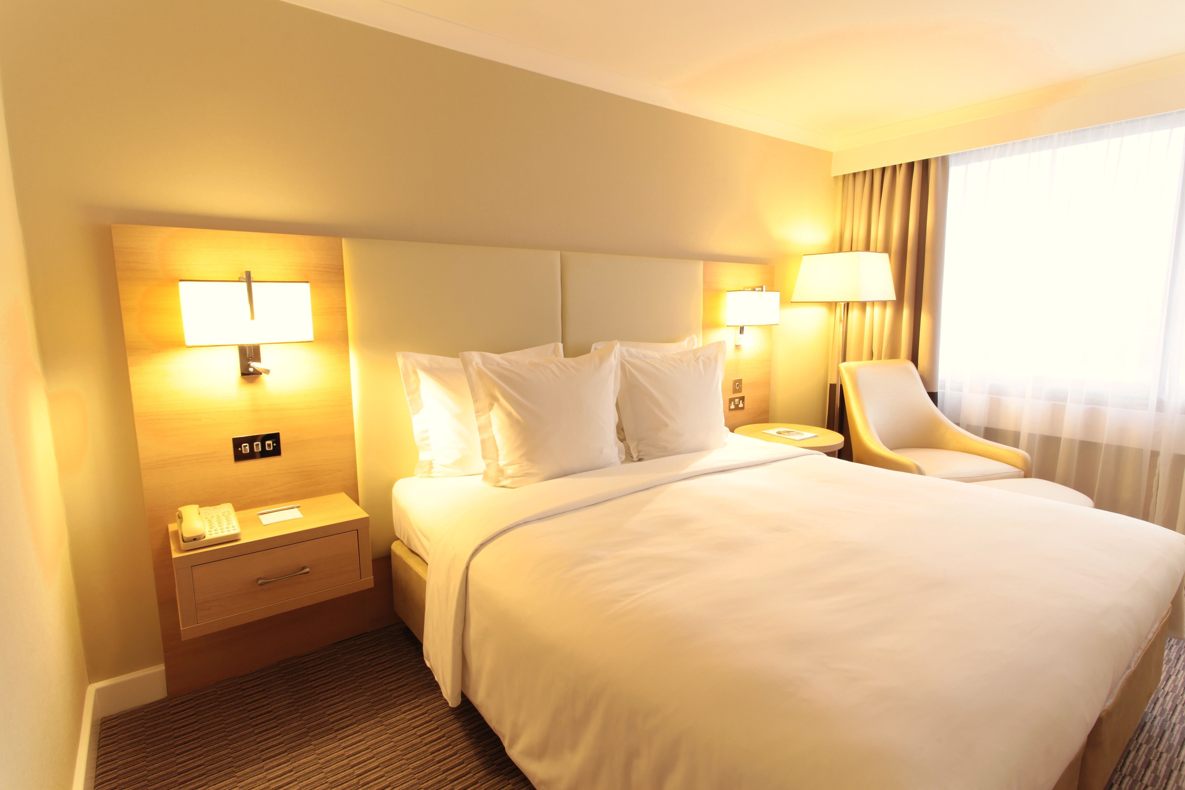 A hotel room with a large bed, two bedside lamps, a chair, a nightstand with a phone, and a window with curtains.