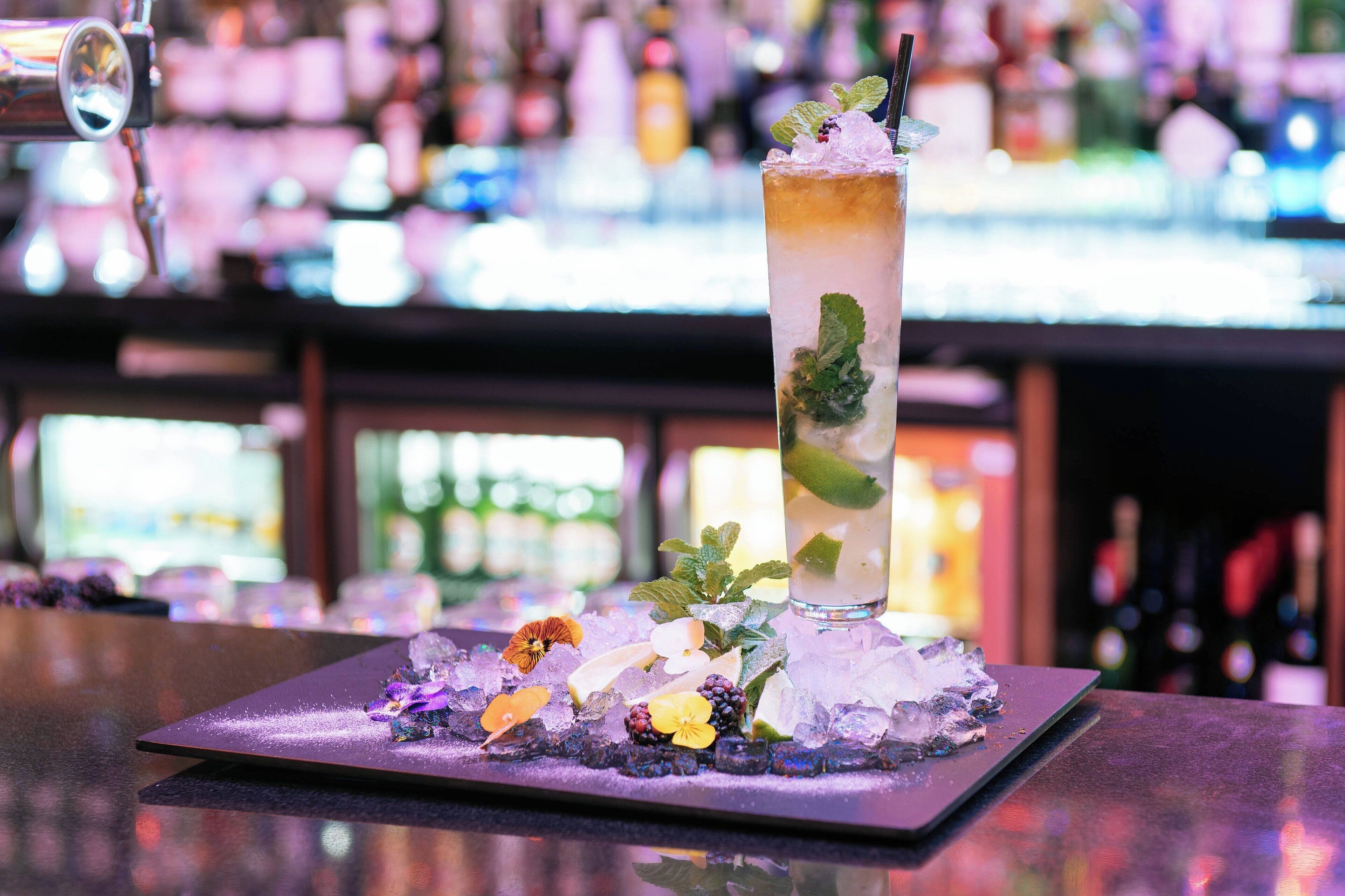 Eine Bar-Szene mit einem hohen Glas mit einem Getränk, garniert mit Minze und Eis, platziert auf einem Tablett mit dekorativen Blumen und Beeren.