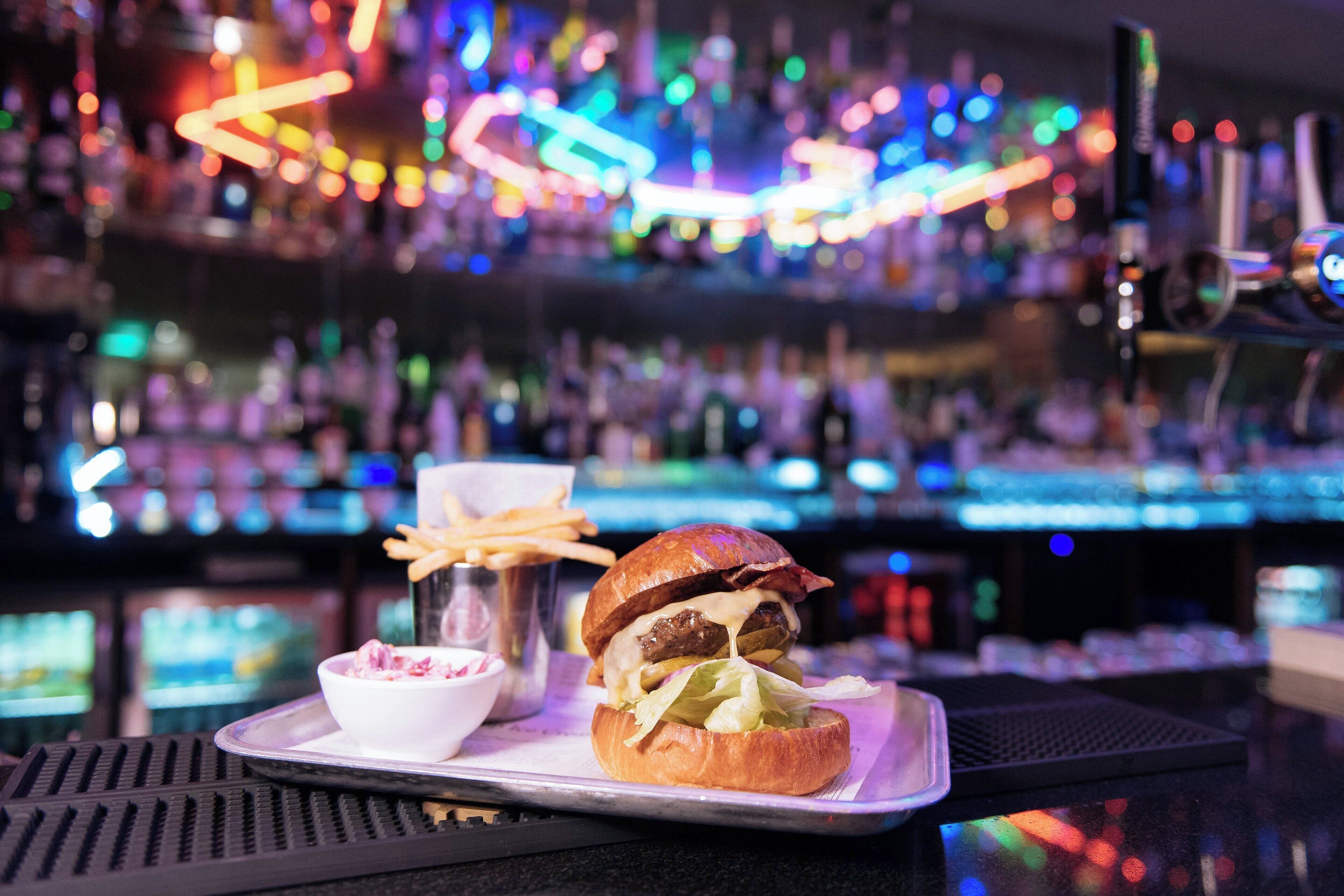 A burger with fries and a small bowl of salad on a tray, set against a bar with neon lights in the background.