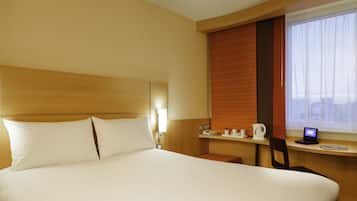 A hotel room with a large bed, a desk with a laptop, a coffee maker, and a window with blinds.