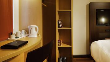 Habitación de hotel con escritorio de madera, una laptop, cafetera, estantería, televisor y cama.