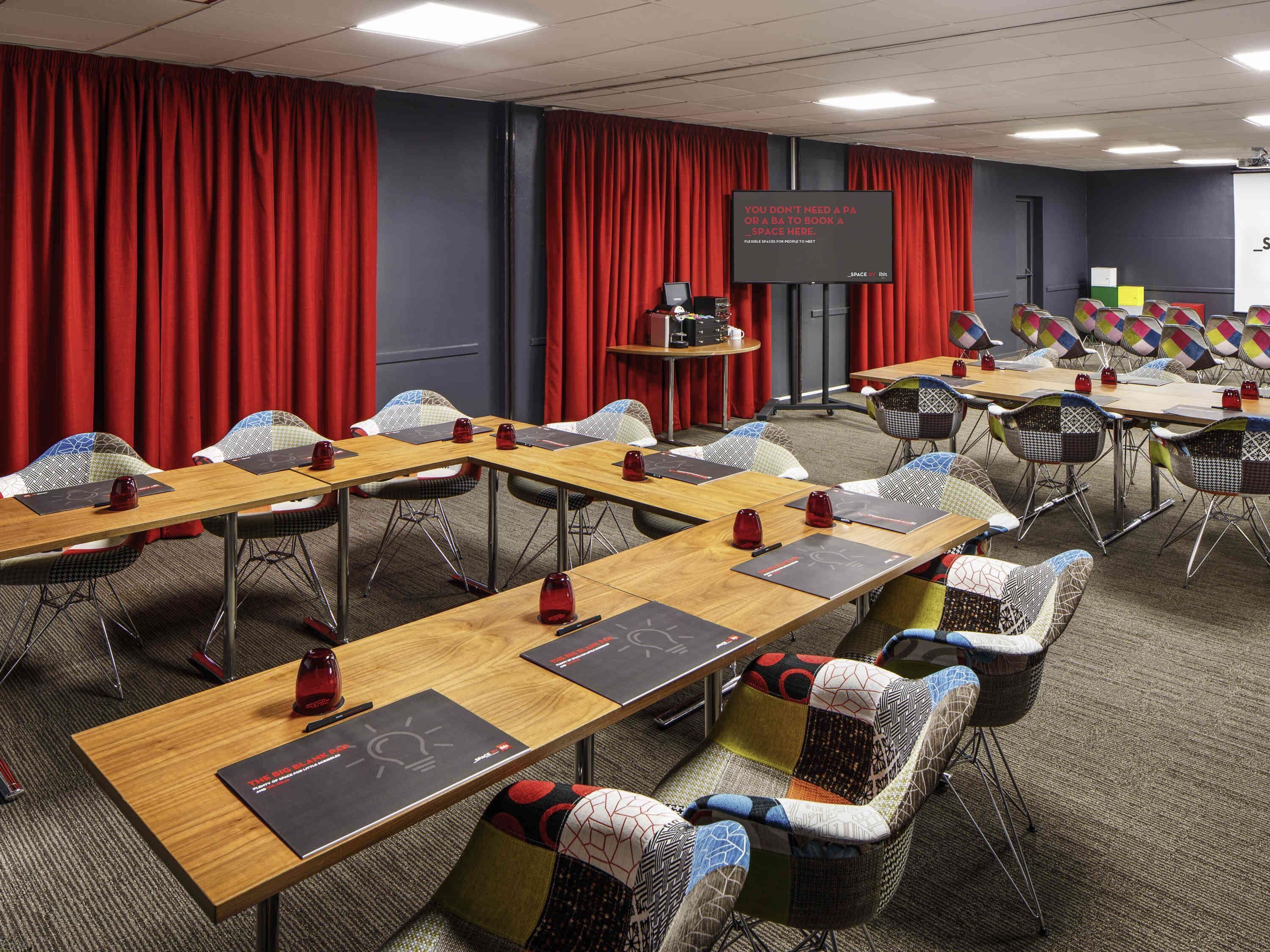 Un salón de conferencias con mesas largas, sillas, una pantalla de proyector y un telón rojo de fondo.
