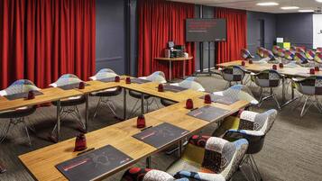 Un salón de conferencias con mesas largas, sillas, una pantalla de proyector y un telón rojo de fondo.