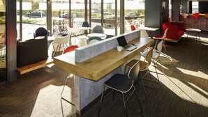 A modern indoor space with a central wooden counter, seating areas with various chairs, and large windows offering views of the outside. - ibis London Luton Airport (Luton)