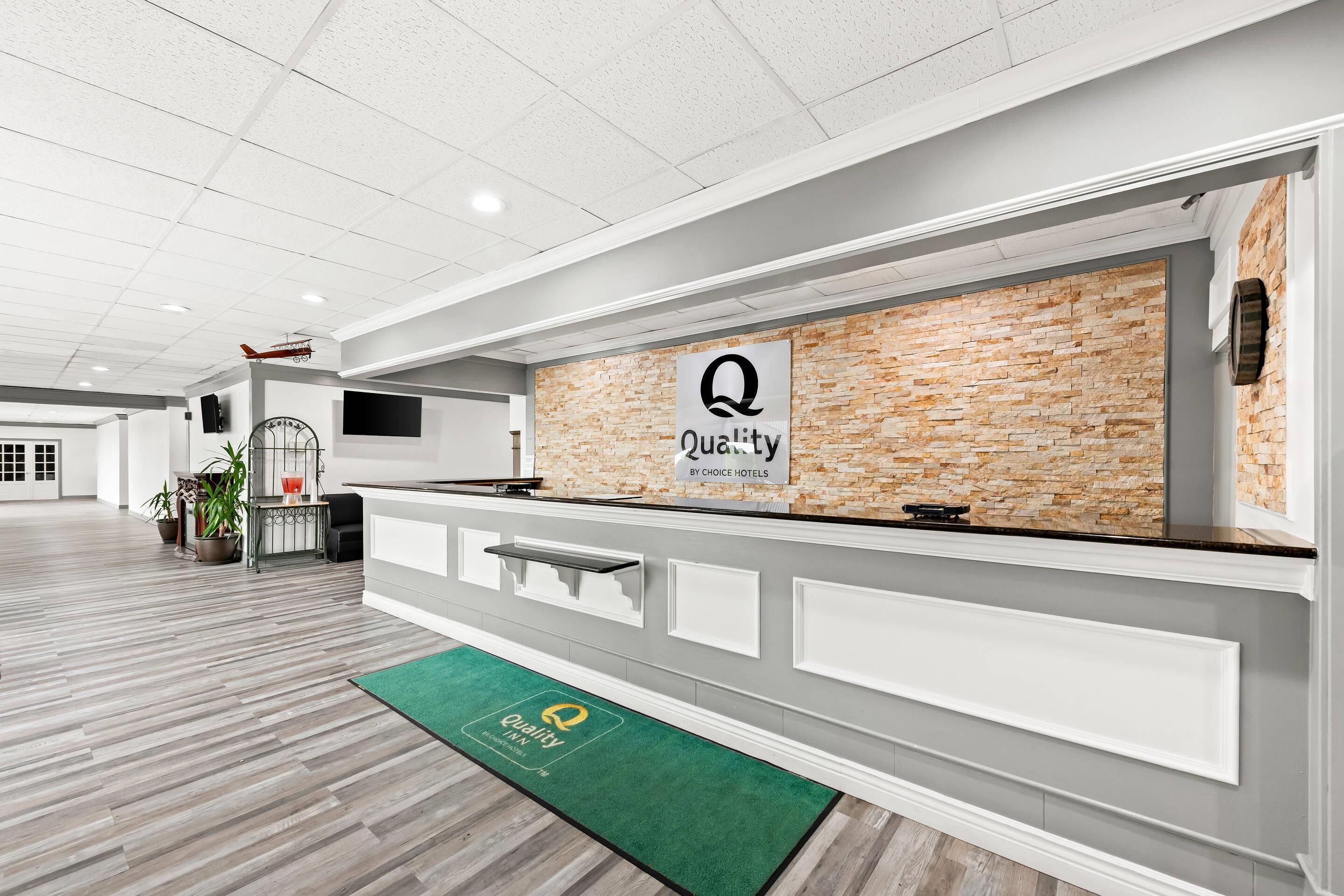 A reception area with a brick wall, a sign reading "Quality by Choice Hotels," a green mat with the hotel logo, and a wooden floor.