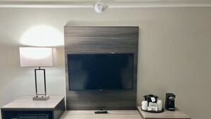 A hotel room with a wooden desk, a TV mounted on the wall, a small refrigerator, a microwave, a coffee maker, and a lamp.