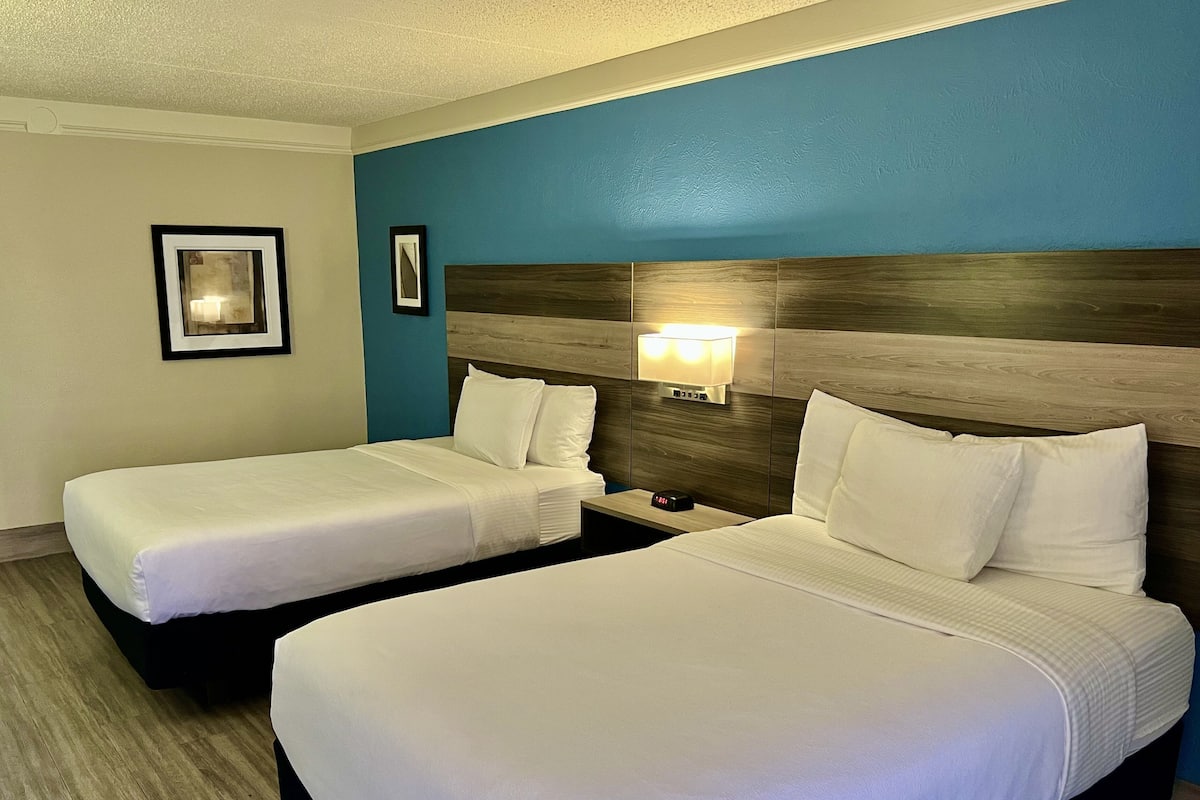 Quarto de hotel com duas camas, uma mesa de cabeceira com abajur e uma luminária na parede.