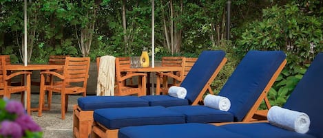 Zona de piscina con sillas de madera y cojines azules, rodeada de vegetación y sombrillas.