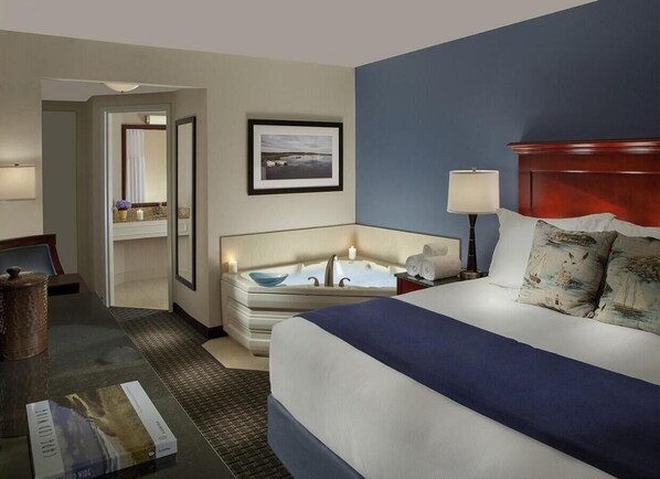 Habitación de hotel con una cama grande, un área de descanso con una silla y una mesa, una bañera y un cuadro enmarcado en la pared.