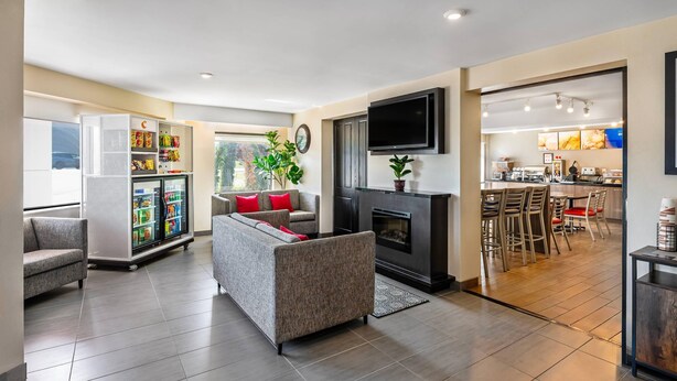 Een moderne woonkamer met een grijze bank, een kleine tafel en een open haard. Een keuken met een bar en krukken is zichtbaar door een open deur.