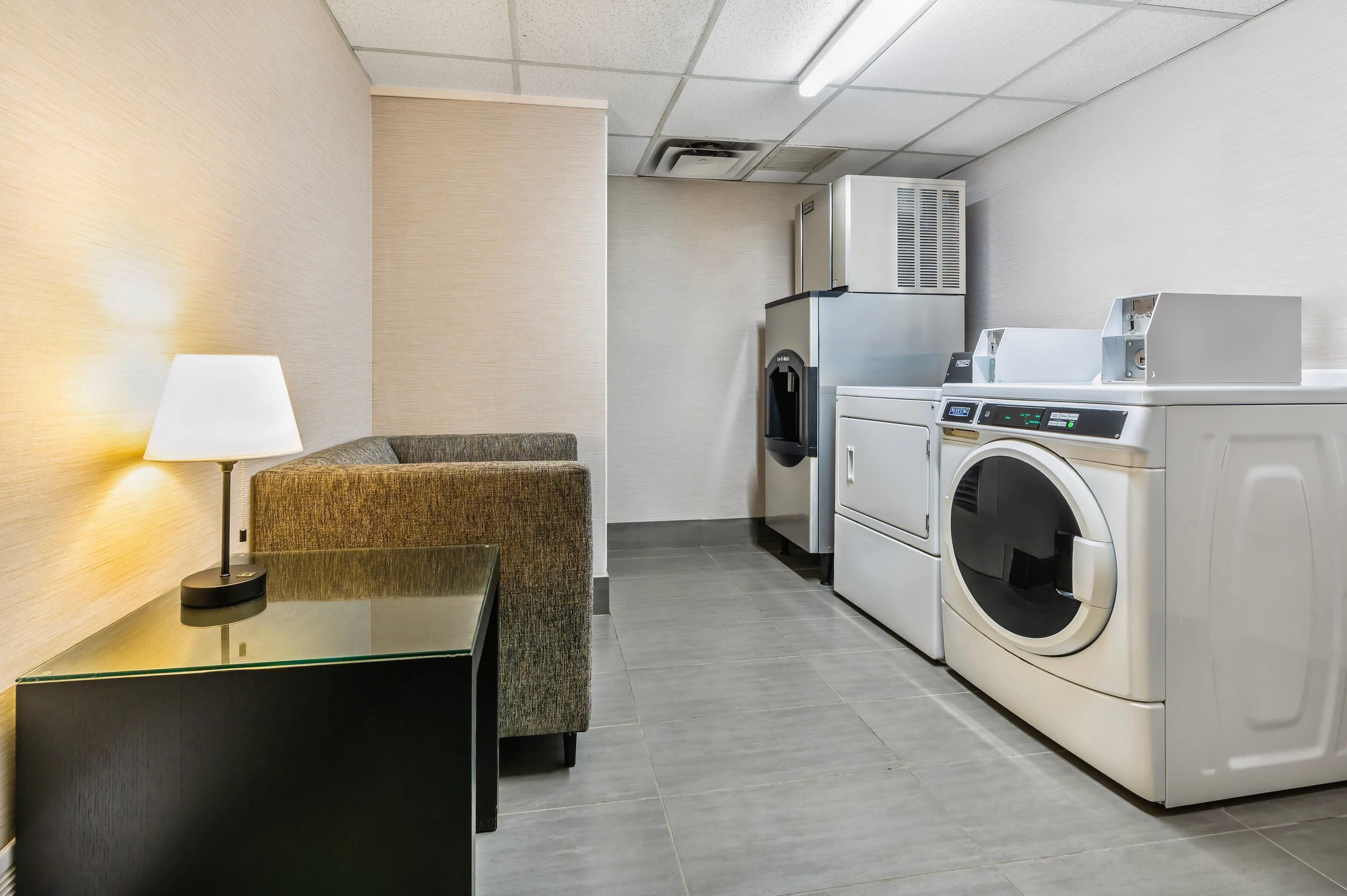 Una habitación de hotel compacta con sofá, mesa con lámpara, lavadora y refrigerador.