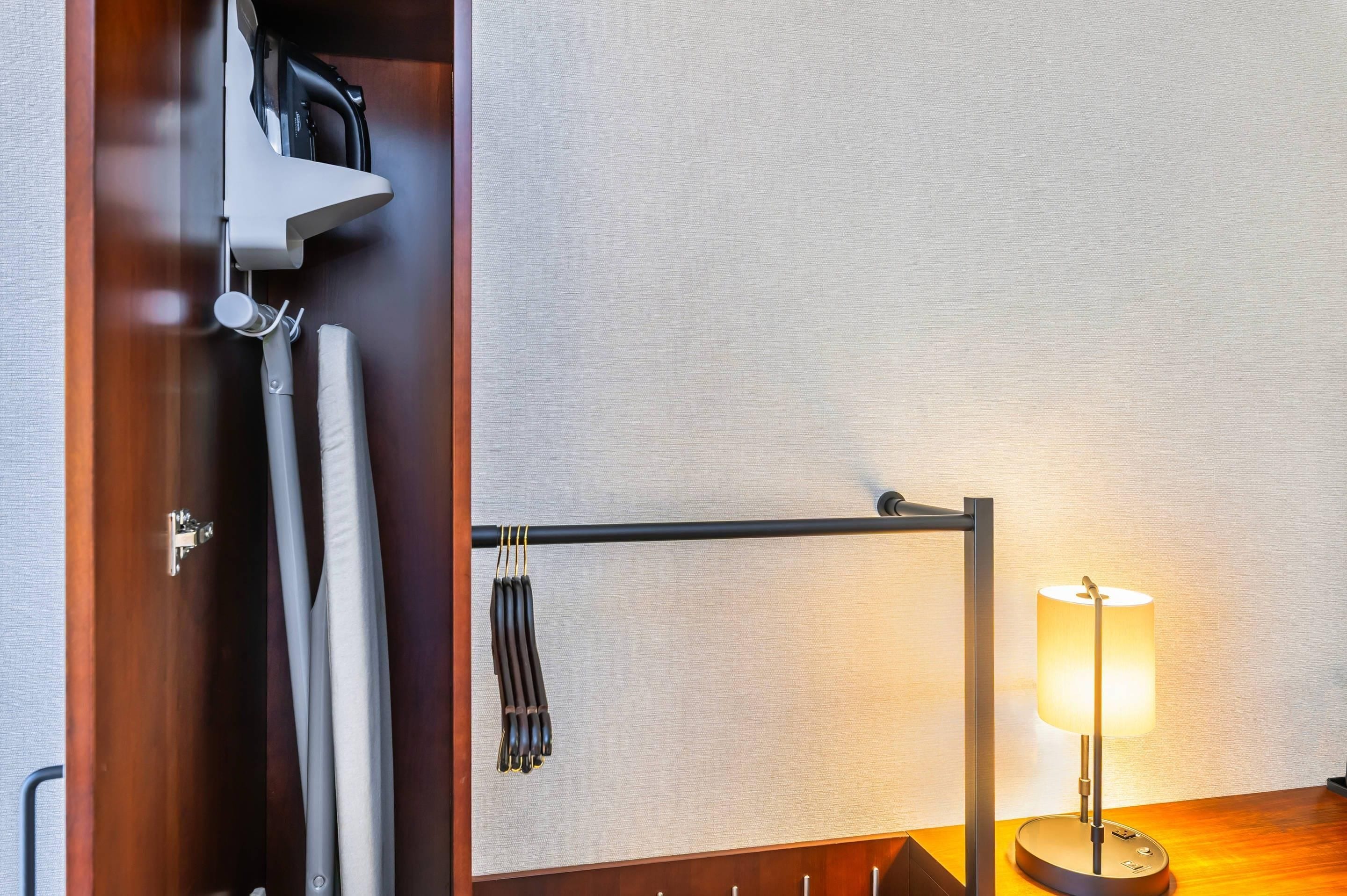 A hotel room with a wooden door, a towel rack with towels, a lamp, and a wall-mounted shelf with a small item.