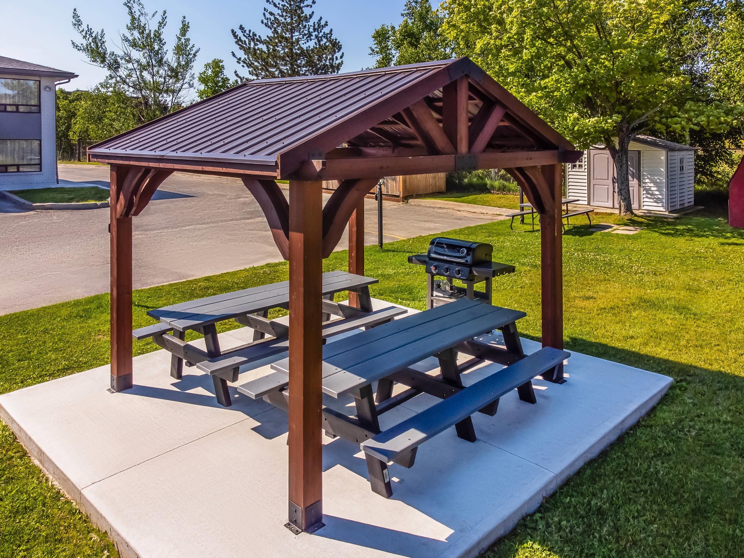 A wooden gazebo with picnic tables, a barbecue grill, and a paved patio area.