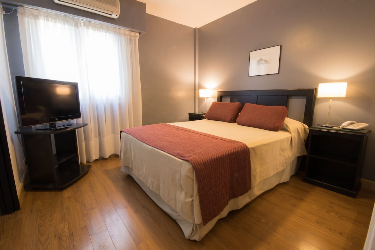 A hotel room with a bed, two bedside tables with lamps, a TV on a stand, and a window with curtains.