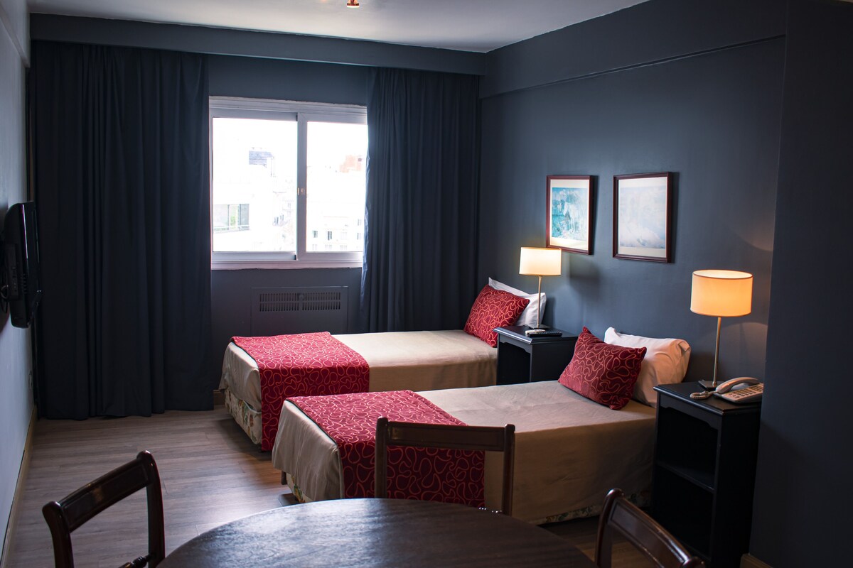 A hotel room with two beds, a table, chairs, a desk with a lamp, a phone, and a window with curtains.