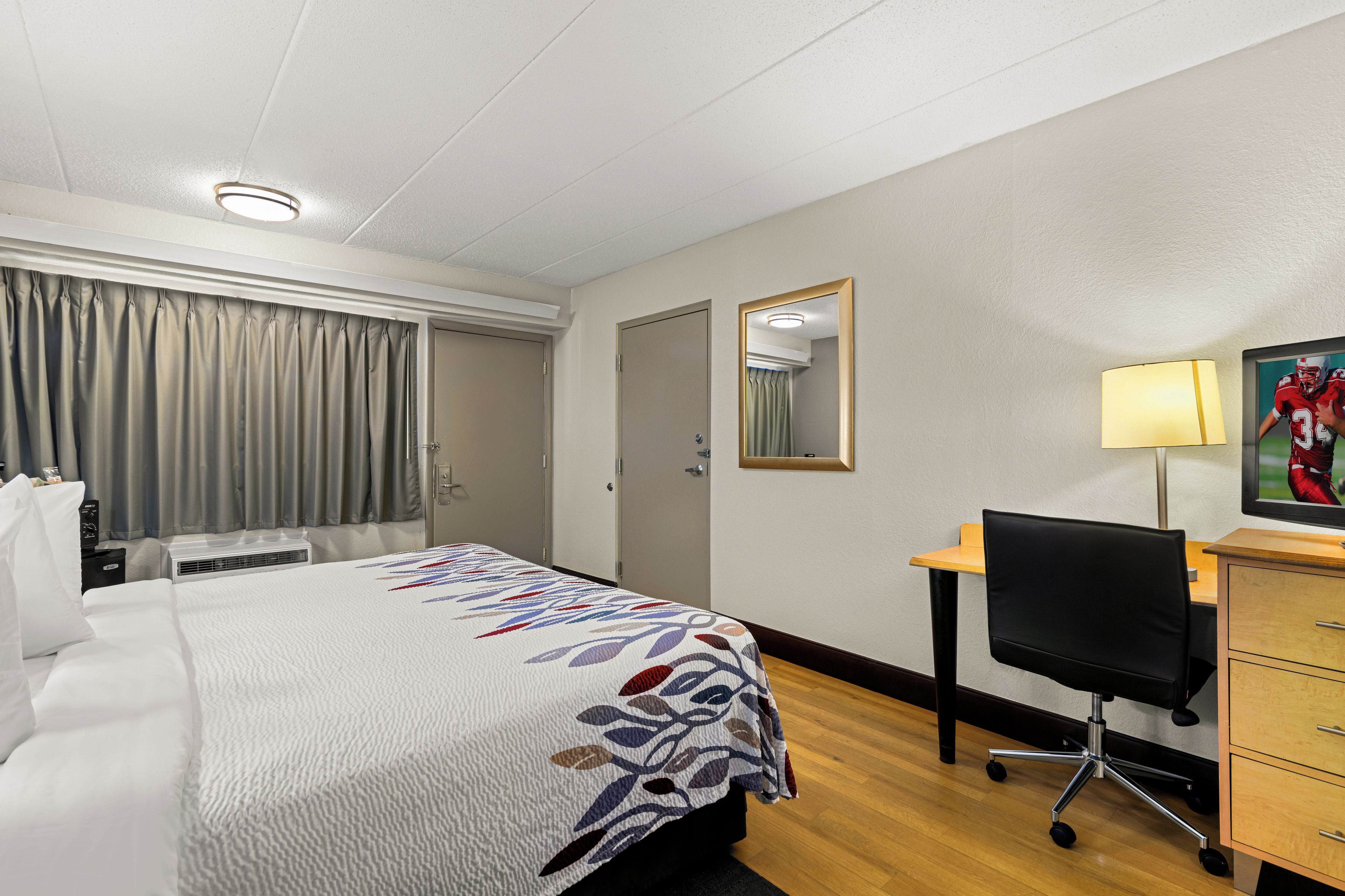 Habitación de hotel con una cama, un escritorio, una silla, una lámpara de escritorio y un espejo.