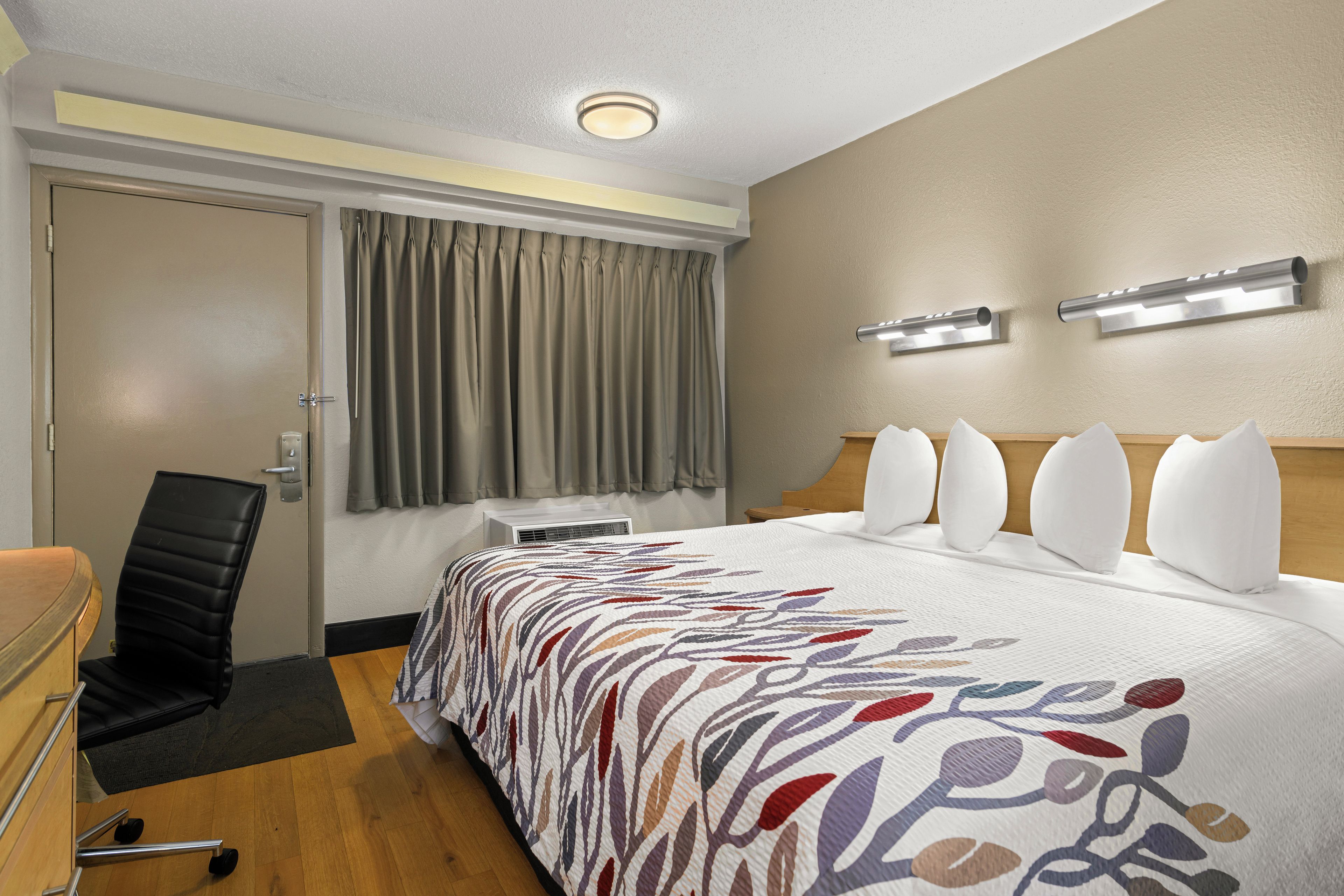 A hotel room with a bed, a desk, an air conditioner, a chair, and a window with curtains.