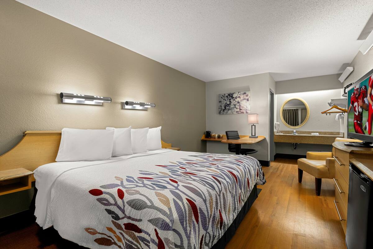 A hotel room with a large bed, a desk with a computer, a chair, a mirror, and a television.