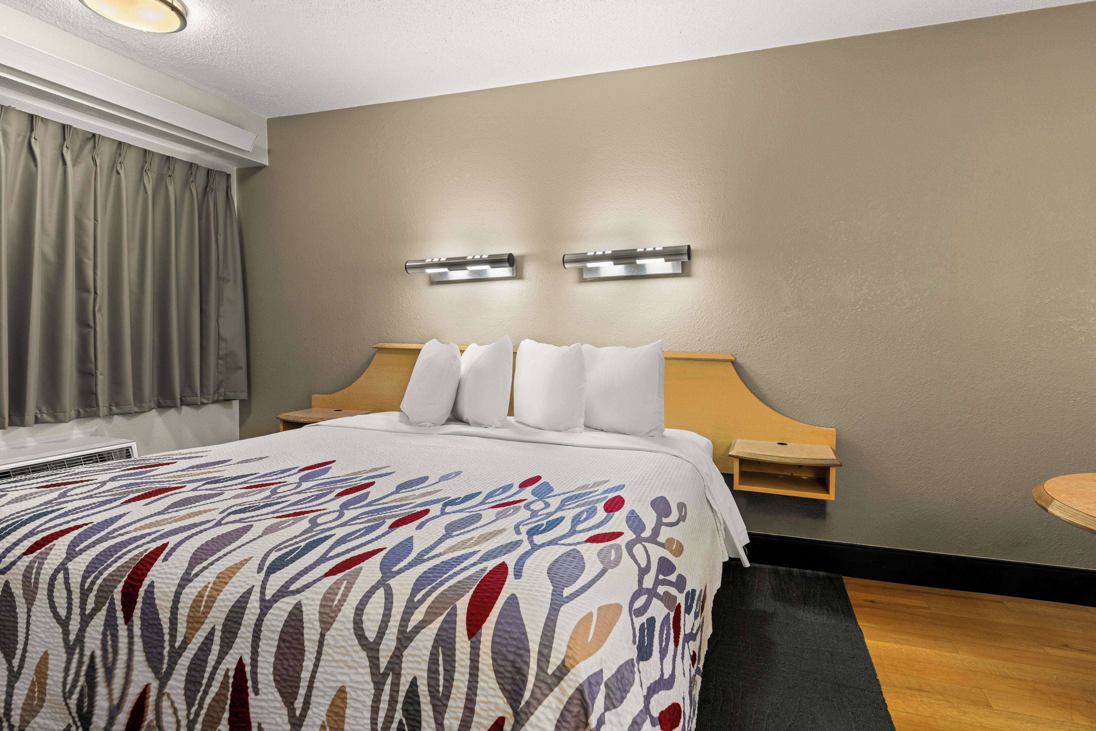 A hotel room with a bed, a nightstand, a wall-mounted light fixture, and a window with curtains.