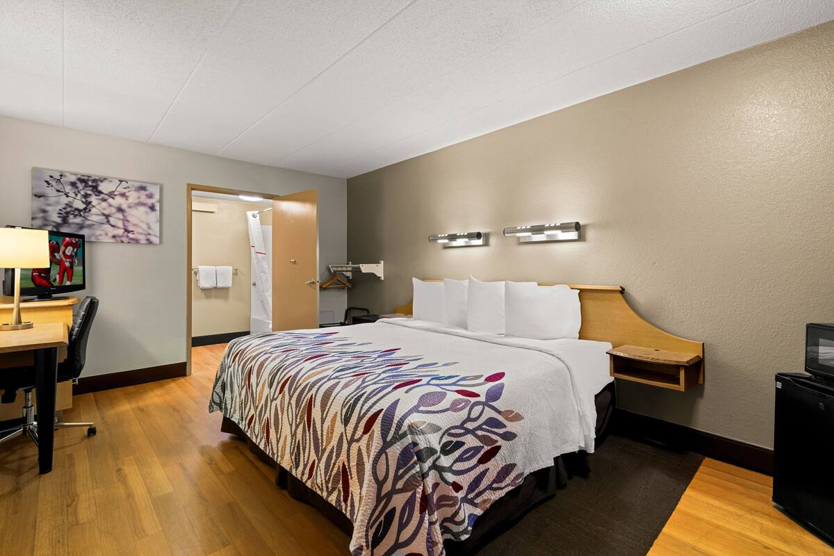 A hotel room with a bed, bedside table, lamp, desk, chair, and a television.