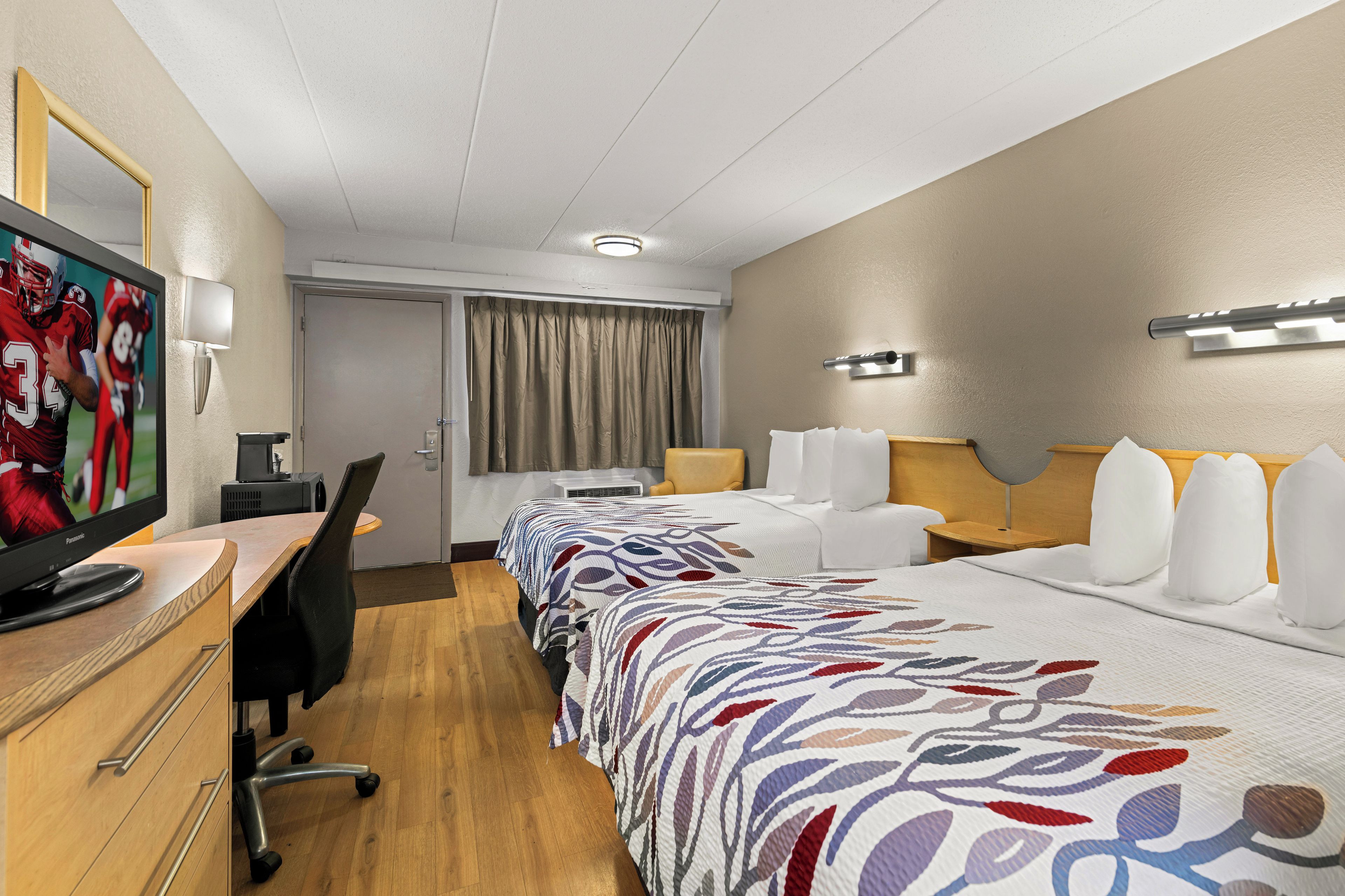 Quarto de hotel com televisão, escrivaninha, cadeira, cama com colcha estampada, mesa de cabeceira com abajur e janela com cortinas.
