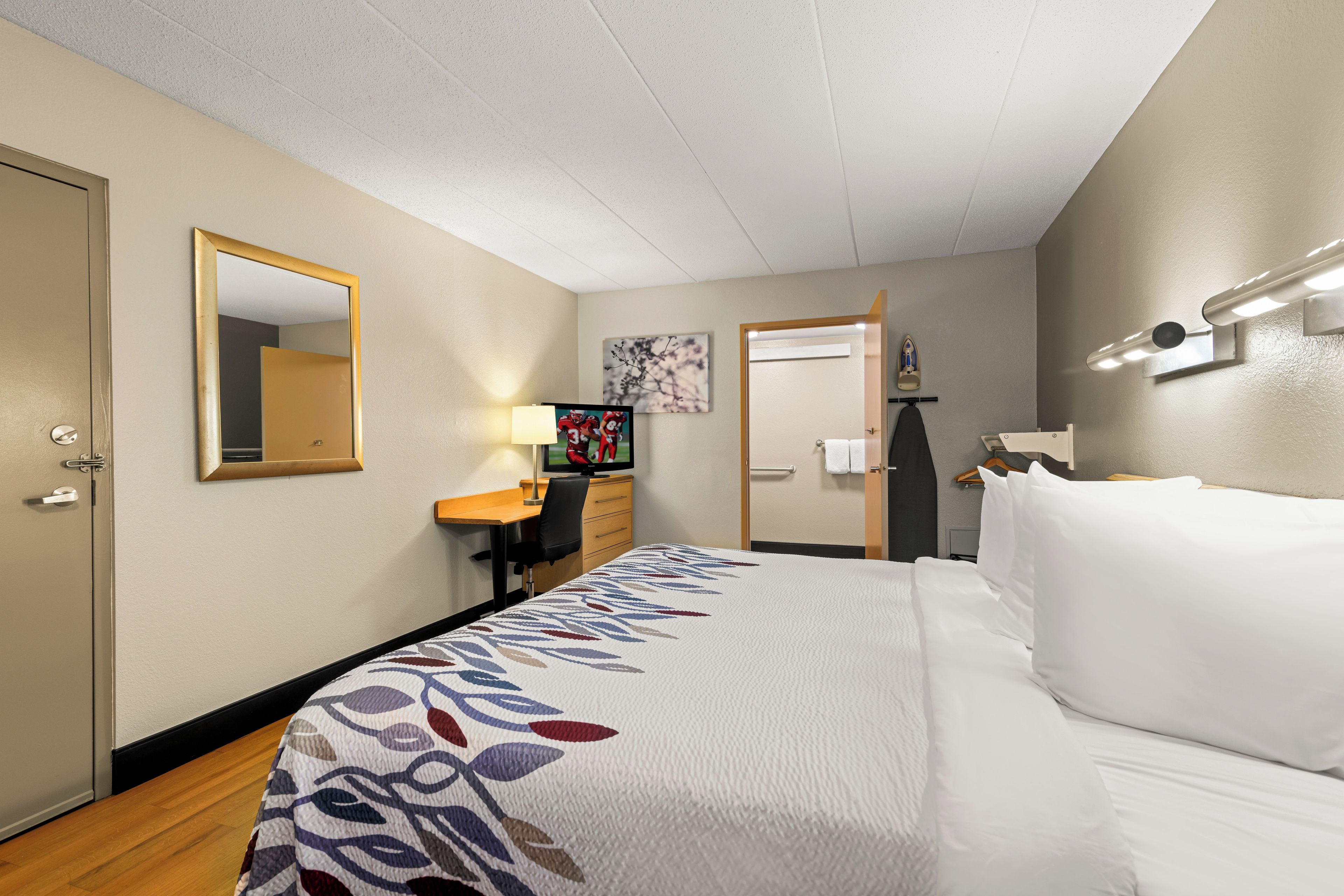 A hotel room with a bed, a desk, a chair, a mirror, a television, and a painting on the wall.